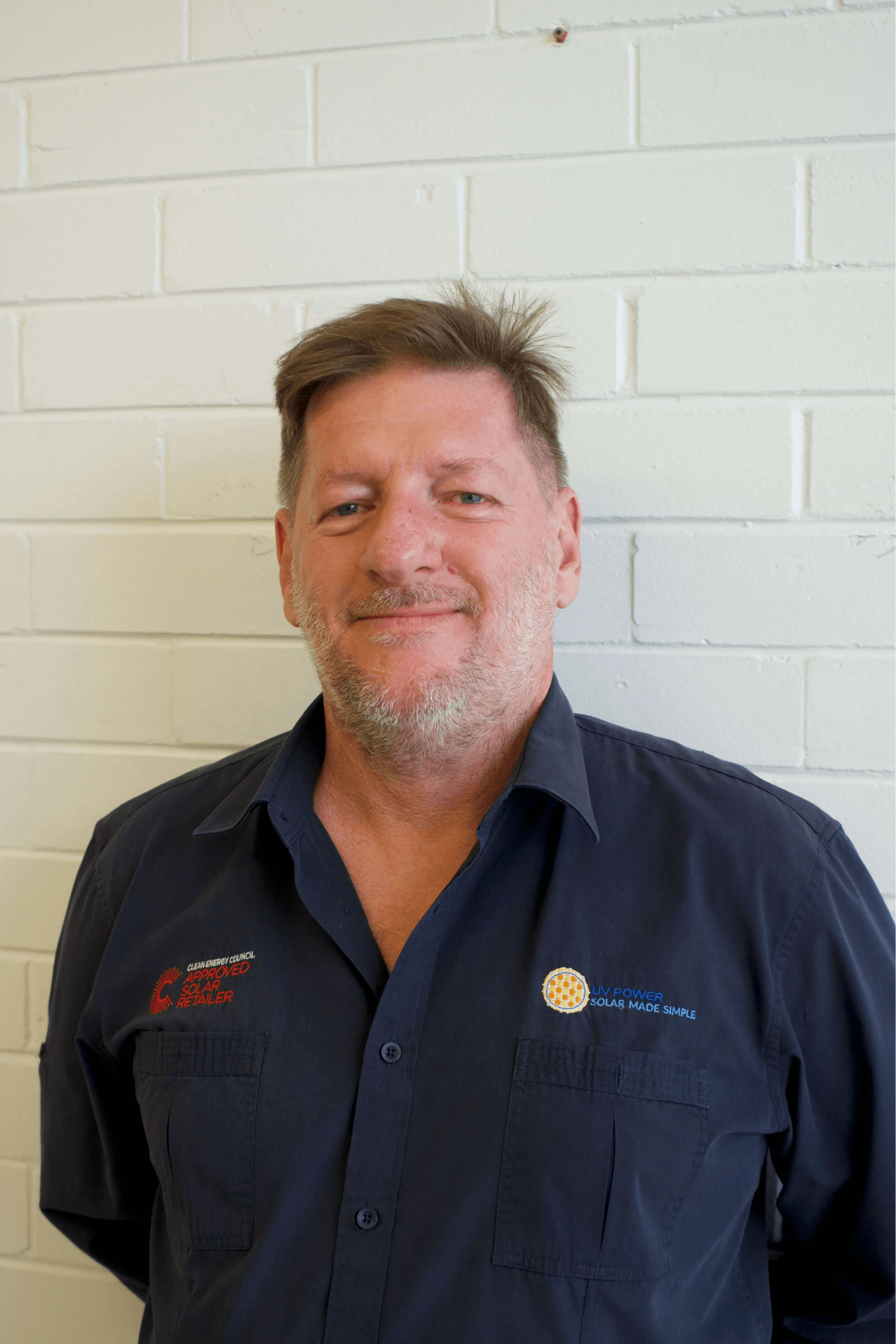 A portrait image of a man - Ian Gittus, the Director of UV Power. Ian is standing in front of a white wall in natural light, wearing a navy UV Power work shirt and is smiling at the camera.