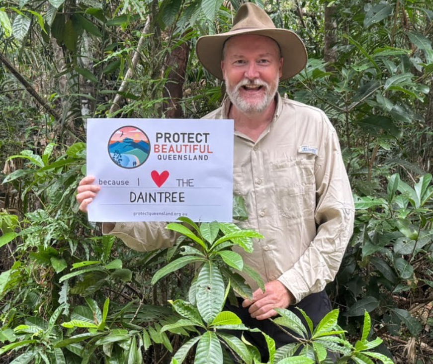 An older male representative of the Save the Daintree program smiling at the camera in the rainforest. The man is holding a sign that says 'Protect Beautiful Queensland. I love the Daintree'.