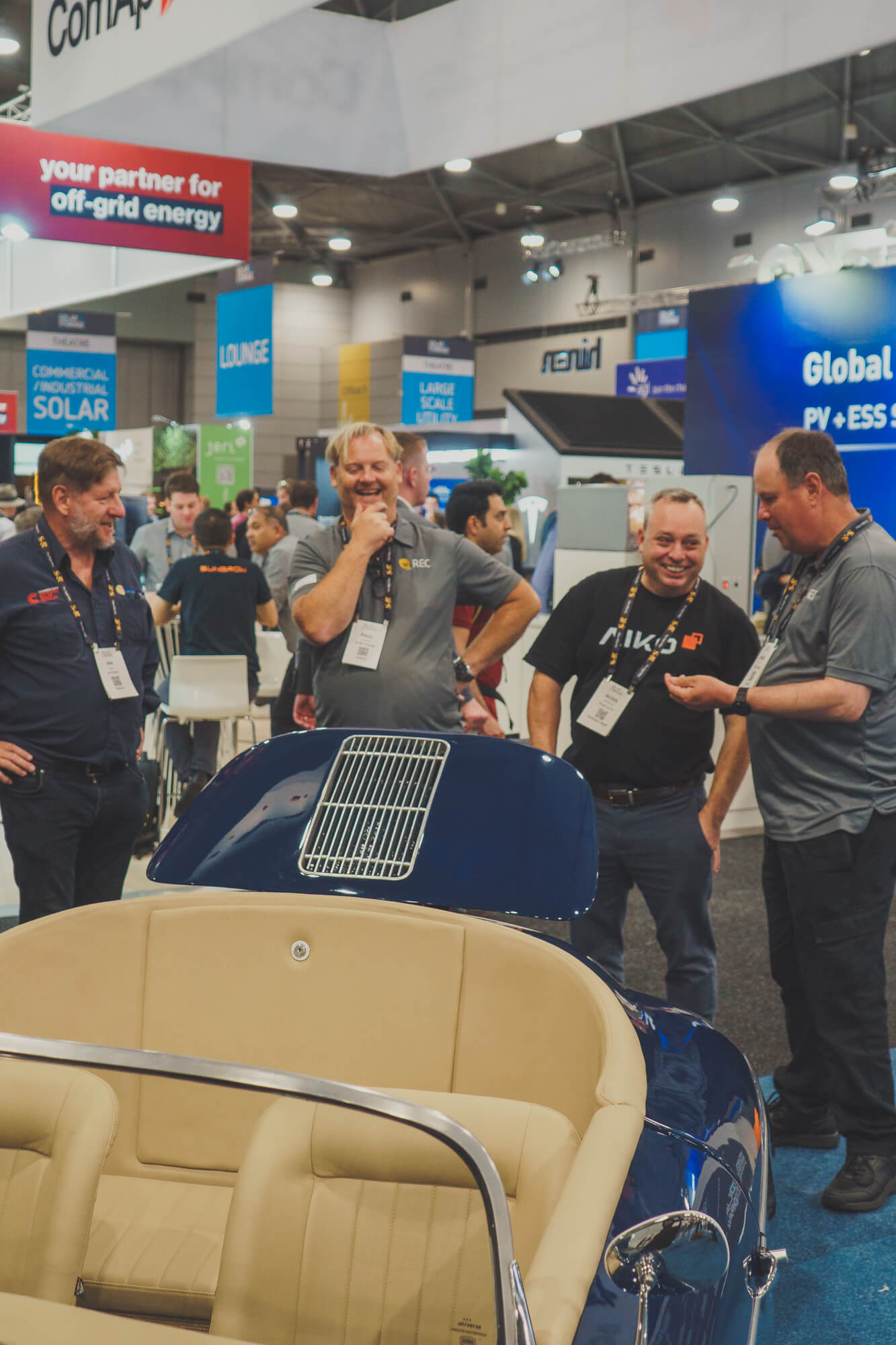 Ian, teh Director of UV Power, with two sales reps from REC Solar Panles, and a rep from AIKO Solar Panles, discussing all things solar as they laugh and joke behind a blue tribute porsche 365 that has been converted into a solar powered vehicle. The vehicle and men are at the Solar Live and Storage Trade Show.