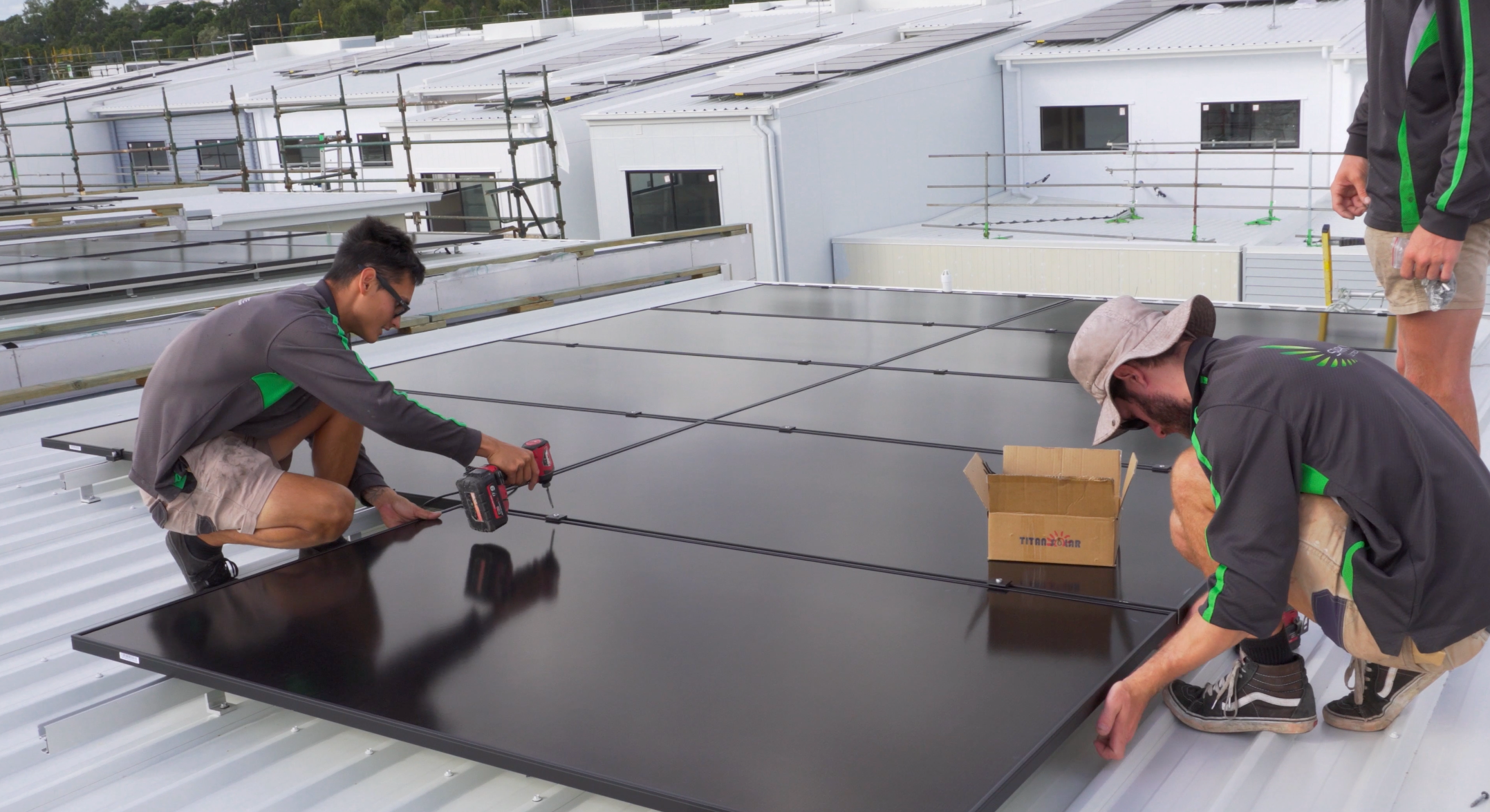 Three solar electricians installing solar panels on a rooftop in Carseldine Village, Brisbane. One installer is using a drew tool, while the other two and on the opposite sides of the panels. One of the other two is standing over the third installer learning how to install the panels.